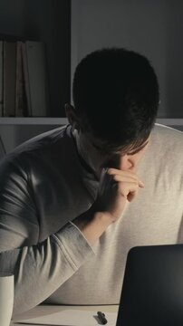 Vertical video. Exhausting job. Internet career. Overwork lifestyle. Sleepy hardworking male freelancer drinking coffee creating project using laptop late in dark home office.