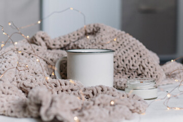 A white ceramic mug and a candle in a glass jar with a wooden wick stand among a knitted plush soft warm plaid. Warm comfort