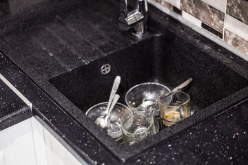 collection of dirty dishes, including glass bowls and small cups, can be seen resting in black sink filled water. remnants meal are visible in the glasses while spoons lie scattered among the items