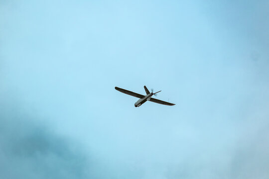 A drone flies against a bright blue sky with scattered clouds, surrounded by blurred green leaves in the foreground. aerial reconnaissance work