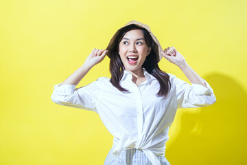 Asian woman wearing a white shirt and striped skirt, playfully holding a beige bucket hat with a cheerful expression against a vibrant yellow background, radiating joy and positivity.