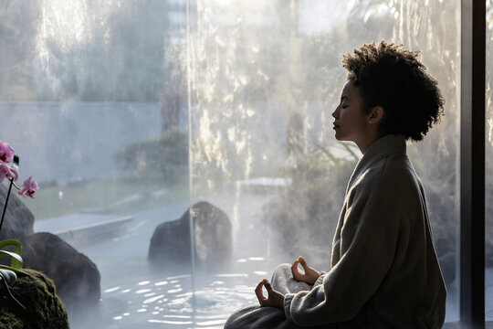 A non-binary individual sits in a plush robe, practicing meditation in a tranquil hotel environment. Soft light illuminates the scene, enhancing the peaceful atmosphere