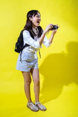 Asian woman with pigtails wearing a white shirt, and a black backpack, holding a camera and smiling enthusiastically, exuding energy and playfulness against a bright yellow background