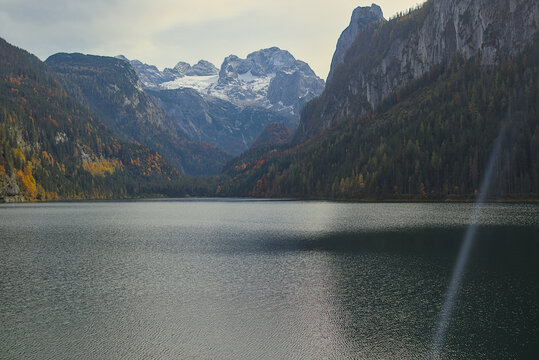 Picturesque Gosausee lake in Austria surrounded by the high snow-capped peaks of the Dachstein Mountains. Dense forests show off the warm hues of autumn. The water reflects the cloudy sky. - Powered by Adobe