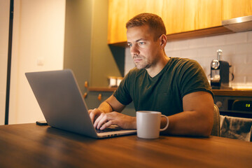 Focused freelancer sitting at kitchen table at cozy home and typing on a laptop.