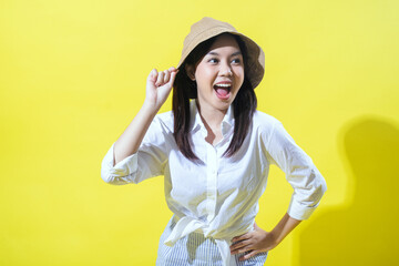 An Asian woman in a white shirt and striped pants poses cheerfully against a yellow background. She wears a beige hat, smiles widely, and has her hand on her hip, exuding vibrant energy.