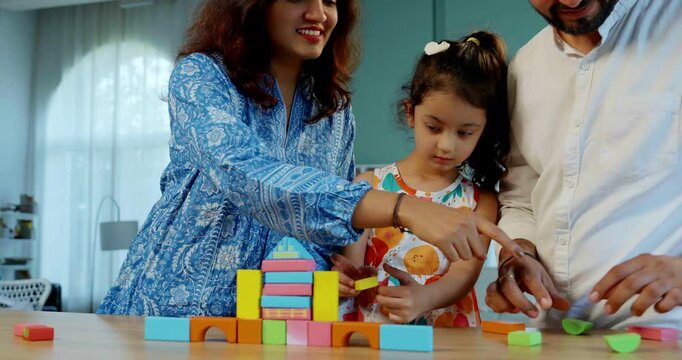 Indian Asian young married couple enjoying quality time with their adorable daughter, building a home using colorful wooden block toys at a table in their modern home