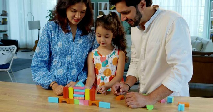 Indian Asian young married couple enjoying quality time with their adorable daughter, building a home using colorful wooden block toys at a table in their modern home