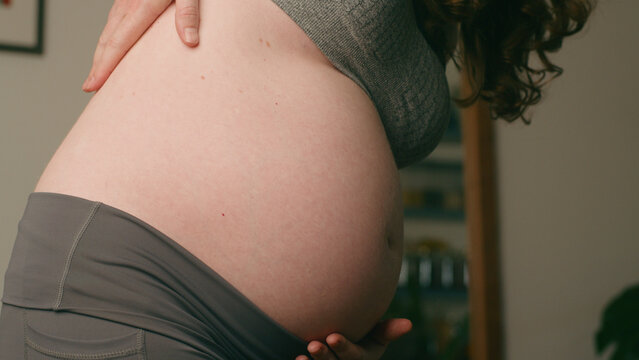 Close-Up View Of A Pregnant Woman's Belly With Hands Cradling It In Warm Home Setting