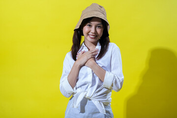 An Asian woman wearing a beige hat and a white shirt with striped pants stands against a yellow background, looking surprised with her hands on her chest and her mouth open in amazement.