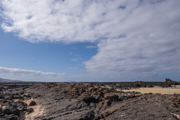 White sand beach with solidified volcanic lava flows, black rocks. Mountains in the background and sky with big white clouds. Caletón Blanco, Lanzarote, Canary Islands, Spain