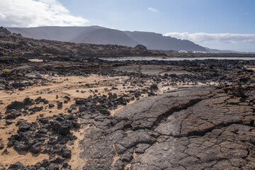 White sand beach with solidified volcanic lava flows, black rocks. Mountains in the background and sky with big white clouds. Caletón Blanco, Lanzarote, Canary Islands, Spain