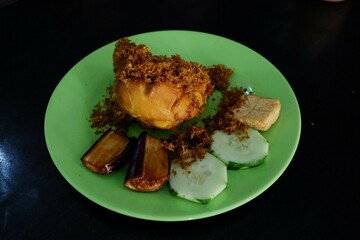 Ayam bakar Penyet is a fried chicken served with hot chili in a plate with fresh vegetables, slices of cucumber and basil leaves