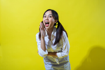 Asian woman making a calling gesture with her hand near her mouth, exuding excitement and energy. She is dressed in a white shirt and striped pants, set against a vibrant yellow background.