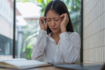 Young Woman in White Shirt Struggling with Stress and Headache While Sitting at a Modern Workspace Surrounded by Greenery and Natural Light