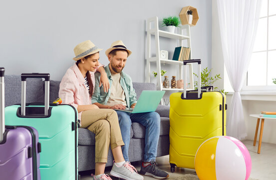Wife and husband, family couple sits on the sofa surrounded by suitcases, preparing for summer vacation. They use a laptop to online book tickets and reserve a hotel, choosing travel destination.
