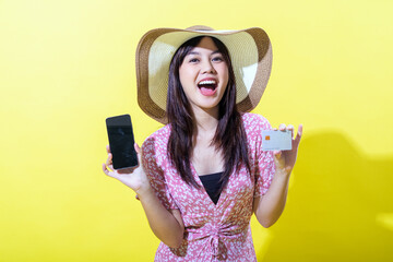 Asian woman in a pink floral dress, wearing a wide-brimmed straw hat, holding a smartphone in one hand and a credit card in the other, smiling brightly against a vibrant yellow background