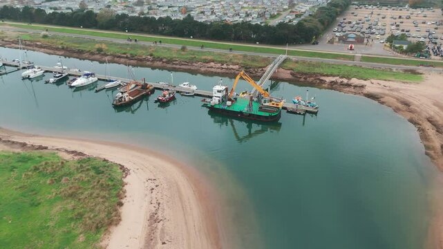 An aerial view of a calm river inlet with docked boats, sandy banks, and a grassy shoreline. A dredging vessel with a crane performs maintenance in the marina near a coastal area.