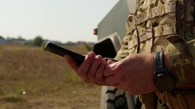 A slow-motion shot of a soldier in camouflage uniform holding a smartphone in a rural area. The video emphasizes military communication and technology during operations in Ukraine