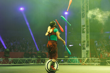 A funny clown on a monocycle juggles glowing pins during a performance in the circus arena.