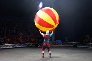 A clown entertains guests in the circus arena in the spotlight, playing with a huge ball. © Studio Peace