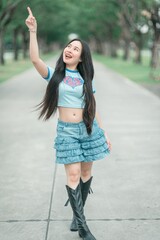 Young Woman in Stylish Outfit with Long Hair Pointing Upward on a Sunny Path Surrounded by Lush Greenery, Evoking a Sense of Joy and Playfulness