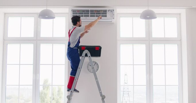Professional repairman from AC maintenance service repairs air conditioner inside house. Young male worker standing on stepladder opens up air conditioner on white wall when his toolbox falls down