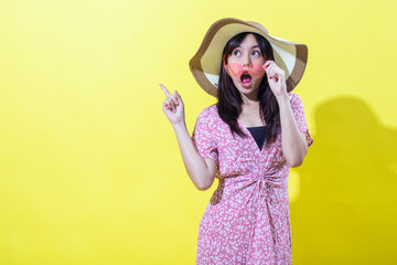 A cheerful woman in a pink floral dress, wearing a wide-brimmed hat, playfully holds pink sunglasses to her mouth while pointing upward, set against a bright yellow background.