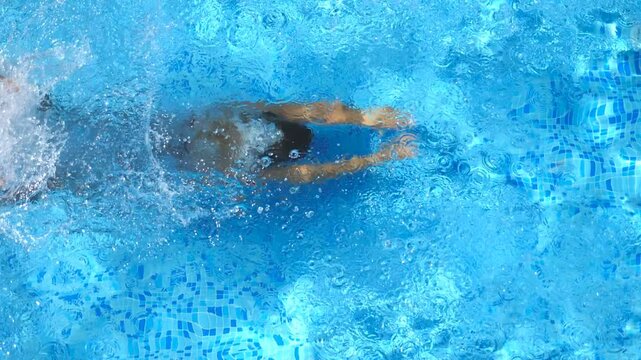 Close up of young tanned woman jumping in pool and splashing crystal clear water. Unrecognizable girl swimming under water in basin of hotel. Lady relaxing during summer vacation. Slow motion Top view