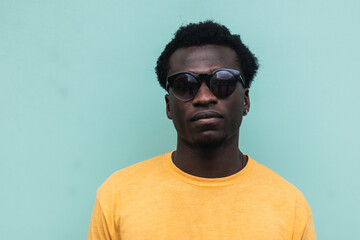 Portrait of a serious young man wearing sunglasses and yellow t-shirt standing against a turquoise background