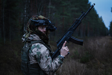 Special forces soldier with an assault rifle in camouflage and with a night vision device in a dark forest.