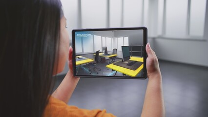 Asian female designer uses tablet computer and virtual reality interior design app to design office workspace. Tablet display shows AI builder software scanning and creating design with 3D furniture.