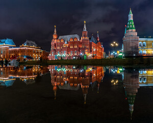 Obraz premium The illuminated State Historical Museum and Kremlin towers reflected in a puddle on a winter night in Moscow, showcasing Russian architecture and the festive atmosphere of Okhotny Ryad