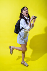 Asian woman with pigtails wearing a white shirt, and a black backpack, holding a camera and smiling enthusiastically, exuding energy and playfulness against a bright yellow background