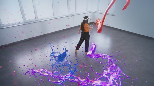 Talented Asian young woman wearing VR headset paints with wireless controllers, uses gestures to create abstract online artwork. Holographic 3D rendering of paint drops. Augmented reality technology.