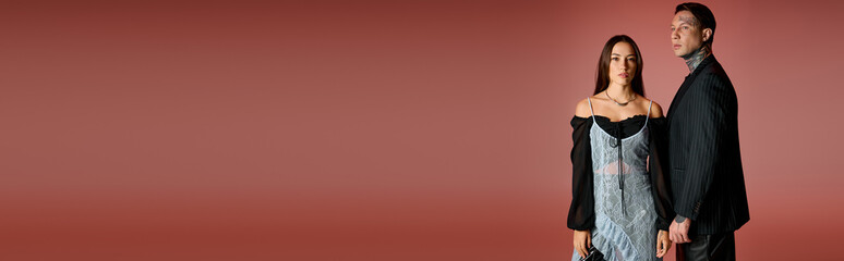 A fashionable couple poses side by side, showcasing their elegant outfits in a warm setting.