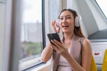 Asian Woman Using Phone and Headphones Listening To Music and Playing Games While Commuting To Work in a Public Train