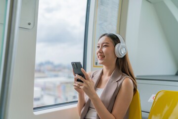 Asian Woman Using Phone and Headphones Listening To Music and Playing Games While Commuting To Work in a Public Train