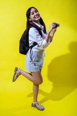 Asian woman with pigtails wearing a white shirt, and a black backpack, holding a camera and smiling enthusiastically, exuding energy and playfulness against a bright yellow background