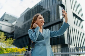 Fototapeta premium Asian Businesswoman Carrying Laptop Bag Using Phone In Front Of A Modern Office Building