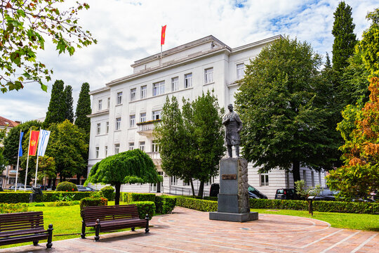 Municipal Assembly of Podgorica, a white building with the Montenegrin flag, trees, and a statue of Marko Miljanov in the foreground, with benches and greenery. Sept 17, 2023. Podgorica, Montenegro