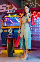 Happy Asian Woman with Shopping Bags Posing in front a Tuk-Tuk with Neon City Lights in a Neon-Lit Studio