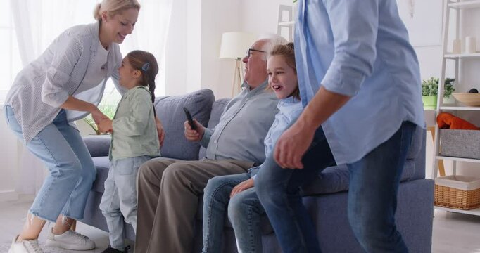 Happy senior aged couple, grandfather, grandmother meet grandson, granddaughter at home watching tv match, comedy movie sitting on cozy couch enjoying free time, three generations weekend together
