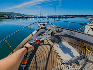 First-person view of a hand holding a hose, spraying water to clean the bow of a boat in sunny...