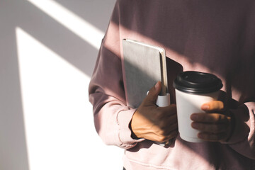 female hand holding a grey diary and white paper cup with hot drink on the background of a pink...
