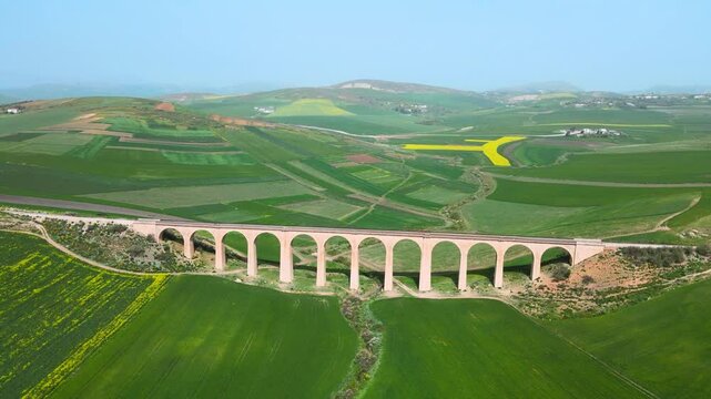 The scene is an aerial view of a bridge crossing over a valley with a train track running through it. On the other side of the valley, there are green fields and mountains in the distance.