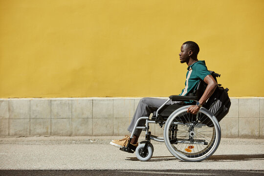 Minimal wide angle shot of African American man with disability using wheelchair in city moving against bright yellow wall with copy space