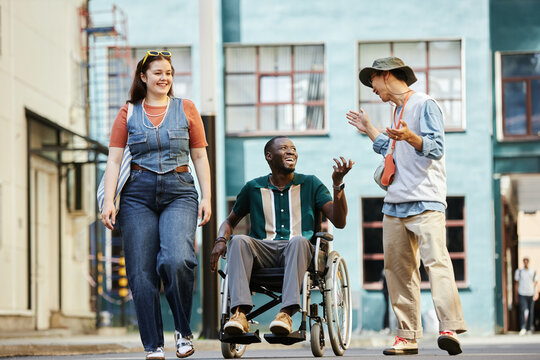 Colorful front view portrait of diverse group of friends with African American man using wheelchair moving towards camera in urban city setting and chatting cheerfully