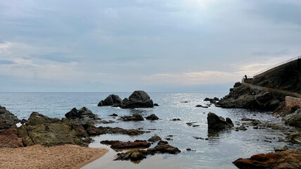 Plage de sable sur la côte espagnole de la Méditerranée au soleil couchant sous un ciel nuageux et orageux d'automne avec des rochers de bord de rivage et un chemin côtier de promenade touristique.