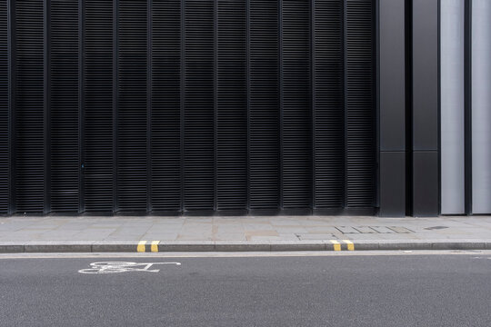 A stark industrial facade characterized by vertical lines and black tones stands next to a road with a clearly marked bicycle path, representing urban modernism and transit.
